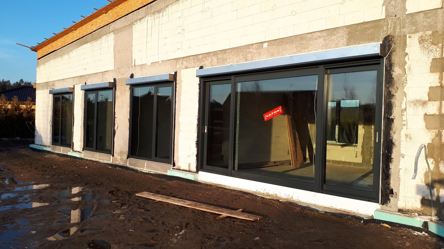 Montaż dużych, przesuwnych okien w surowym stanie budynku; widoczne niedokończone elewacje i błoto na zewnątrz, okna z naklejoną czerwoną etykietą.