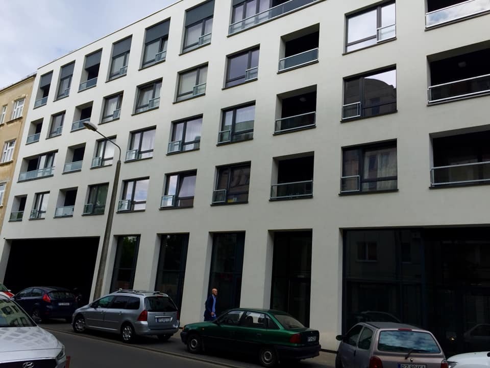 Nowoczesny budynek biurowy z licznymi oknami o regularnym układzie, niektóre z małymi balkonami, widok od dołu z ulicy z zaparkowanymi samochodami.