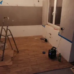 Wnętrze w trakcie prac: ułożona podłoga z paneli imitujących drewno, drabina malarska, ściany częściowo pomalowane, zabezpieczone taśmą malarską, widoczny sprzęt budowlany i butelka wody.