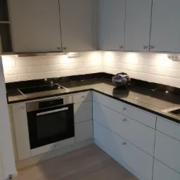 Nowoczesna kuchnia z szarymi szafkami, czarnym blatem i białymi płytkami metra, oświetlona punktowymi lampami podszafkowymi. Na blacie stoi niebieski hełm budowlany.