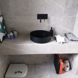 Nowoczesna łazienka z szarą, marmurkową glazurą, czarną umywalką nablatową i baterią podtynkową, z widoczną klapą rewizyjną i narzędziami pod blatem.