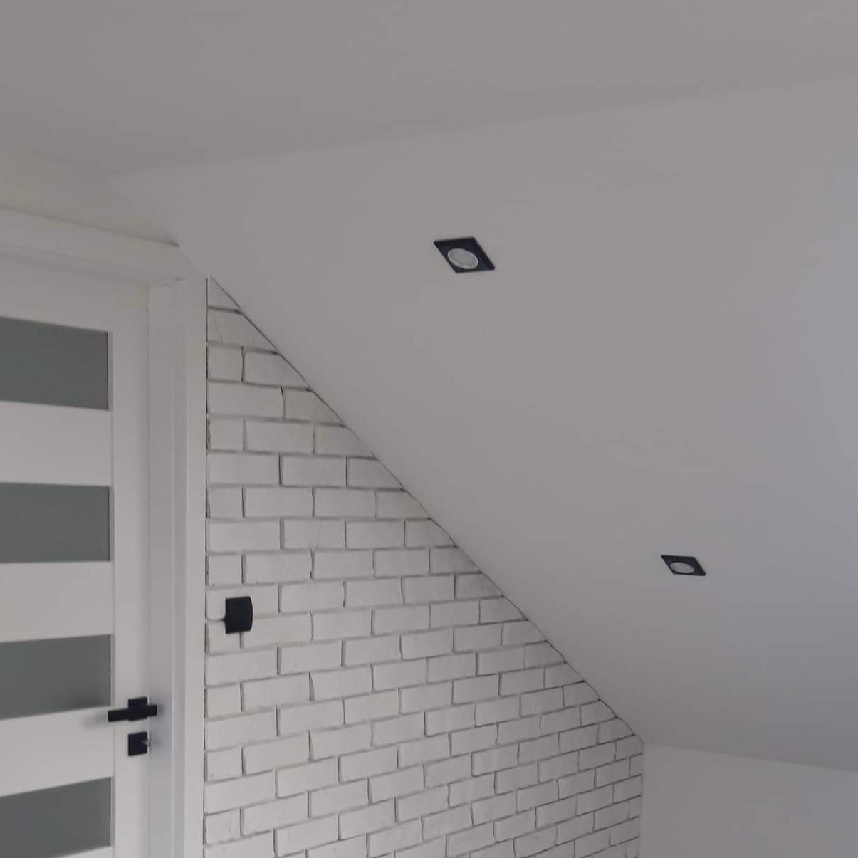 Biała ściana wyłożona cegłą w skosie poddasza, sąsiadująca z białą ścianą i drzwiami z przeszkleniami, z zamontowanymi czarnymi, kwadratowymi lampami punktowymi.