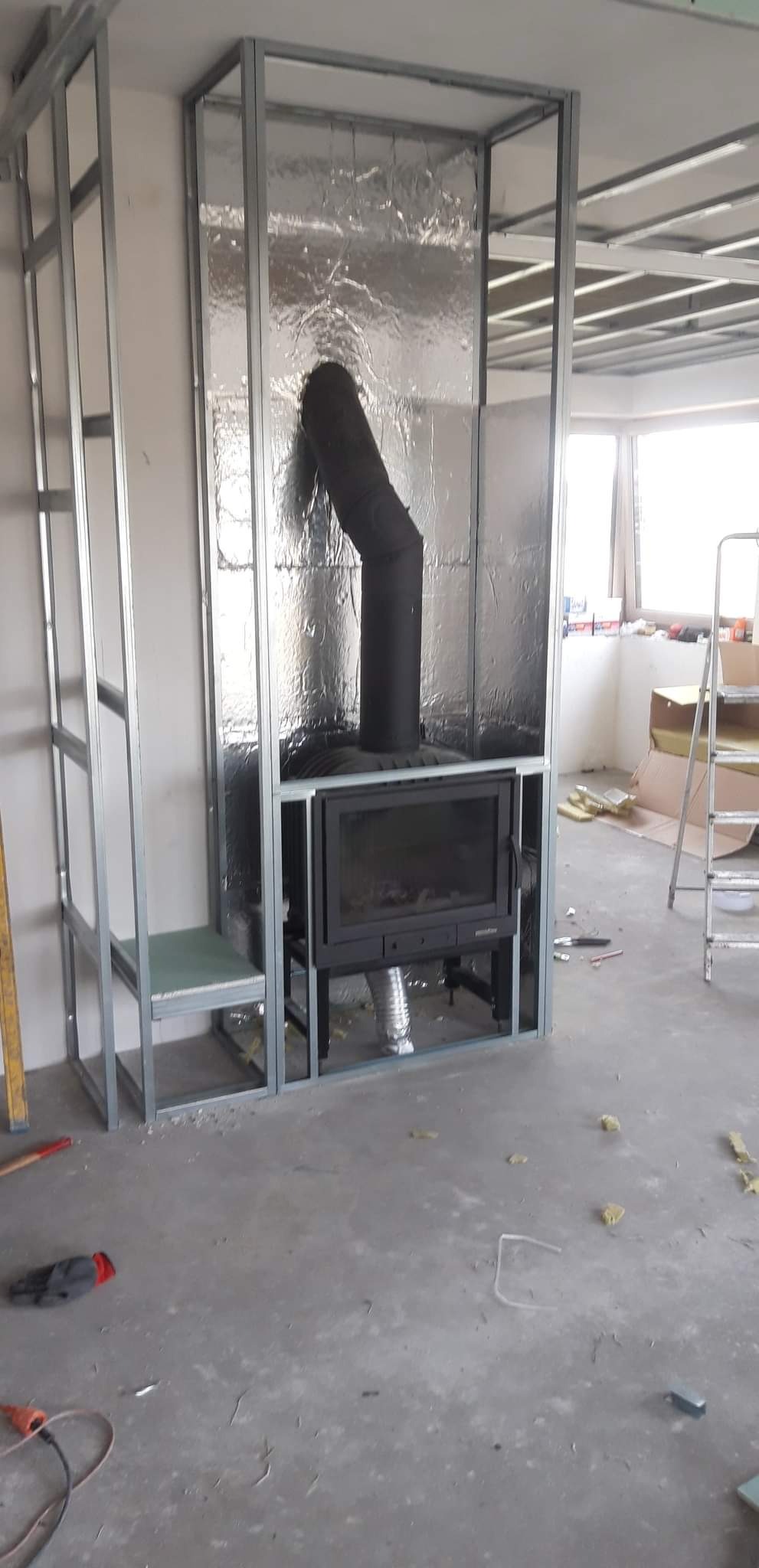 Montaż kominka w trakcie zabudowy: widoczne metalowe profile, izolacja termiczna i podłączenie rury dymowej, surowy beton na podłodze.