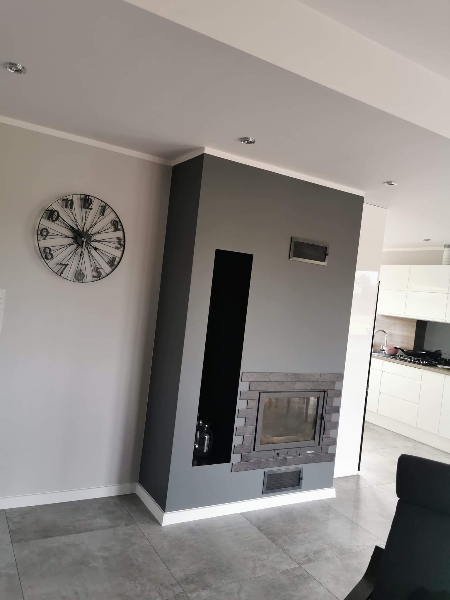 Szary kominek z obudową z cegieł, wnęką i kratką wentylacyjną, na tle szarej ściany z zegarem i fragmentem białej kuchni, na podłodze szare płytki