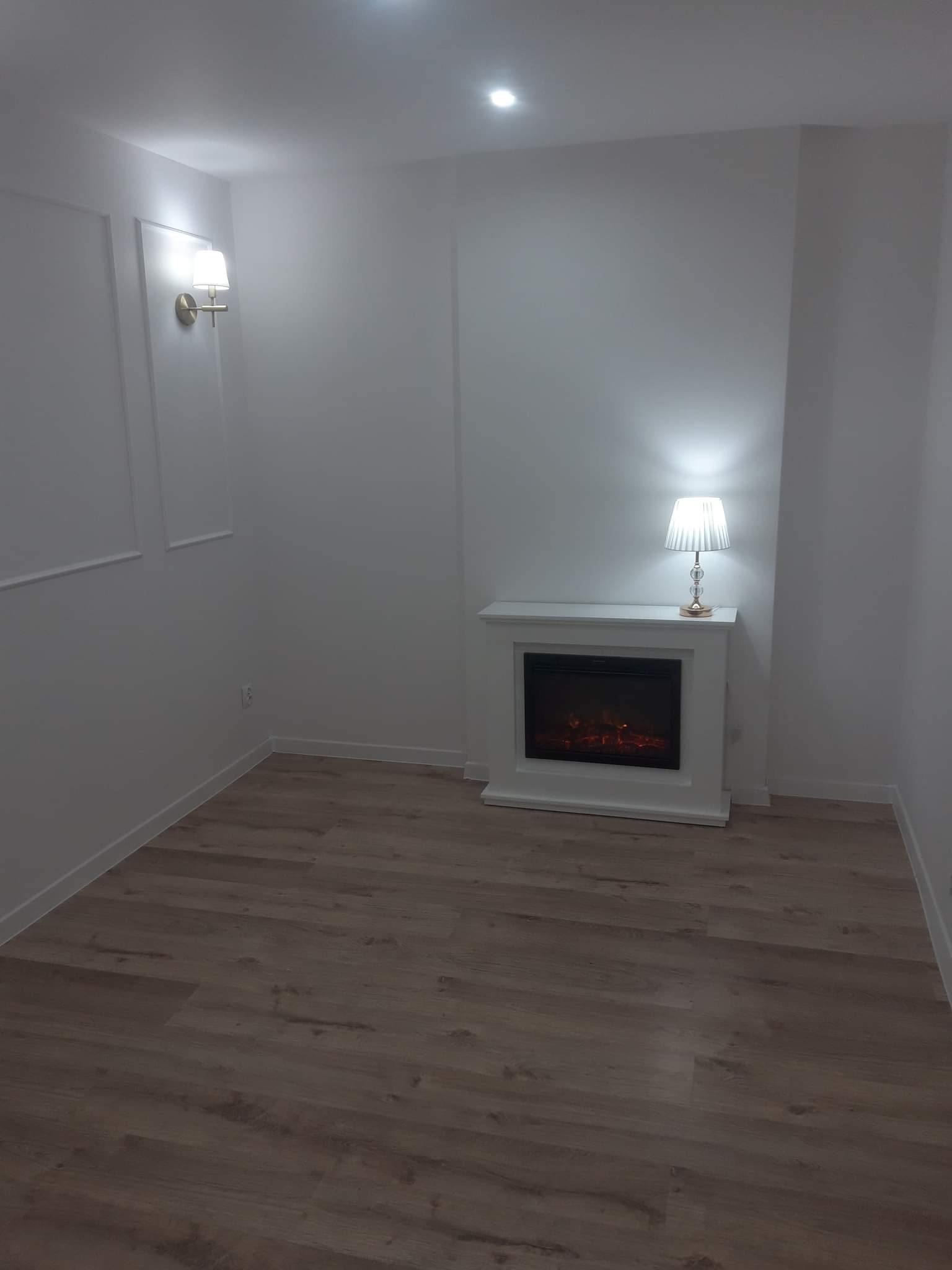 Minimalistyczne wnętrze z białymi ścianami, panelami podłogowymi w odcieniach brązu, elektrycznym kominkiem z lampką i złotym kinkietem.