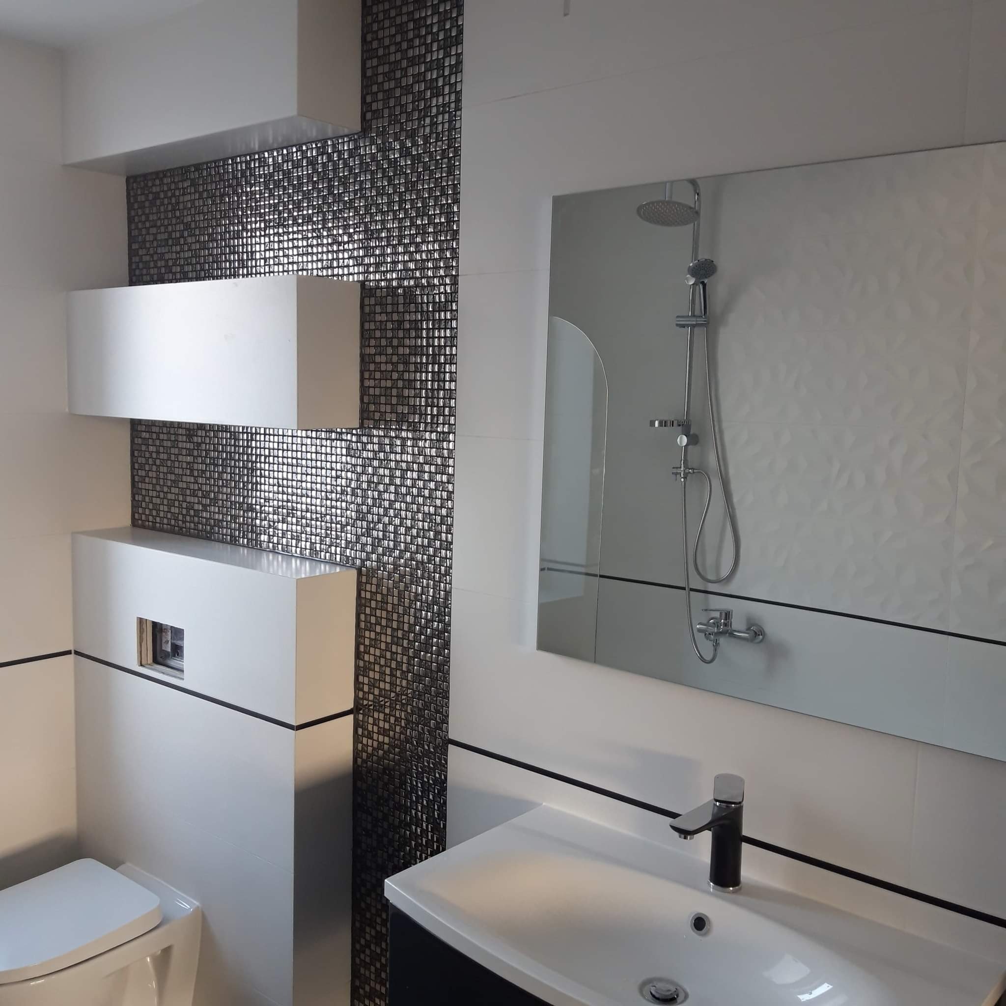 Nowoczesna łazienka z błyszczącą mozaiką w odcieniach szarości, podwieszaną toaletą z zabudową, umywalką z czarną armaturą i dużym lustrem odbijającym kabinę prysznicową. Minimalistyczny design...