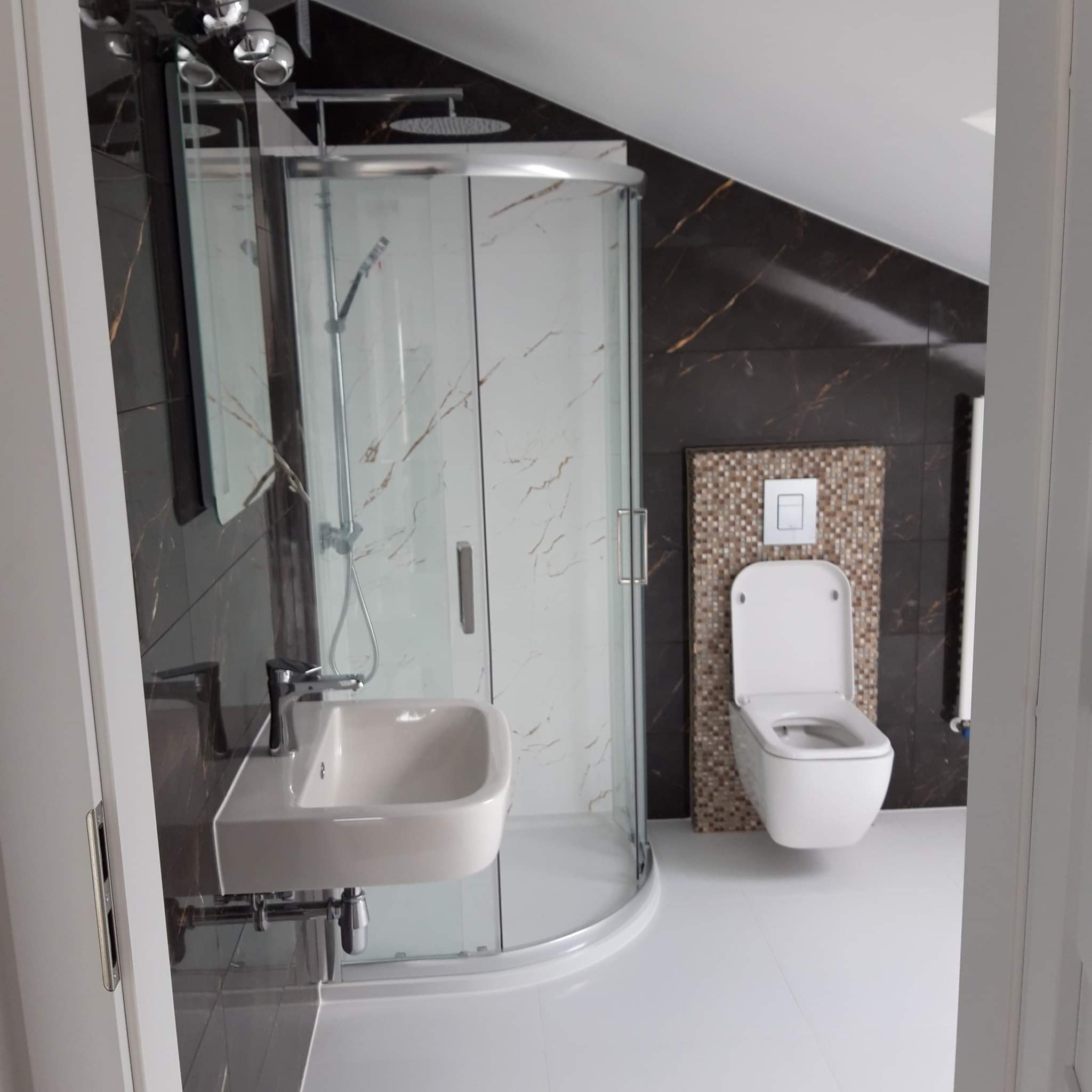 Nowoczesna łazienka na poddaszu z kabiną prysznicową z białym marmurem, wiszącą umywalką i toaletą na tle ciemnych płytek z żyłkami i mozaiki, biała podłoga i skośny sufit.