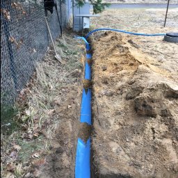 Łukasz polis - Wykop z niebieską rurą w ziemi, widoczny fragment instalacji przy ogrodzeniu z siatki. Szpadel wbity w ziemię obok wykopu. Etap prac instalacyjnych.