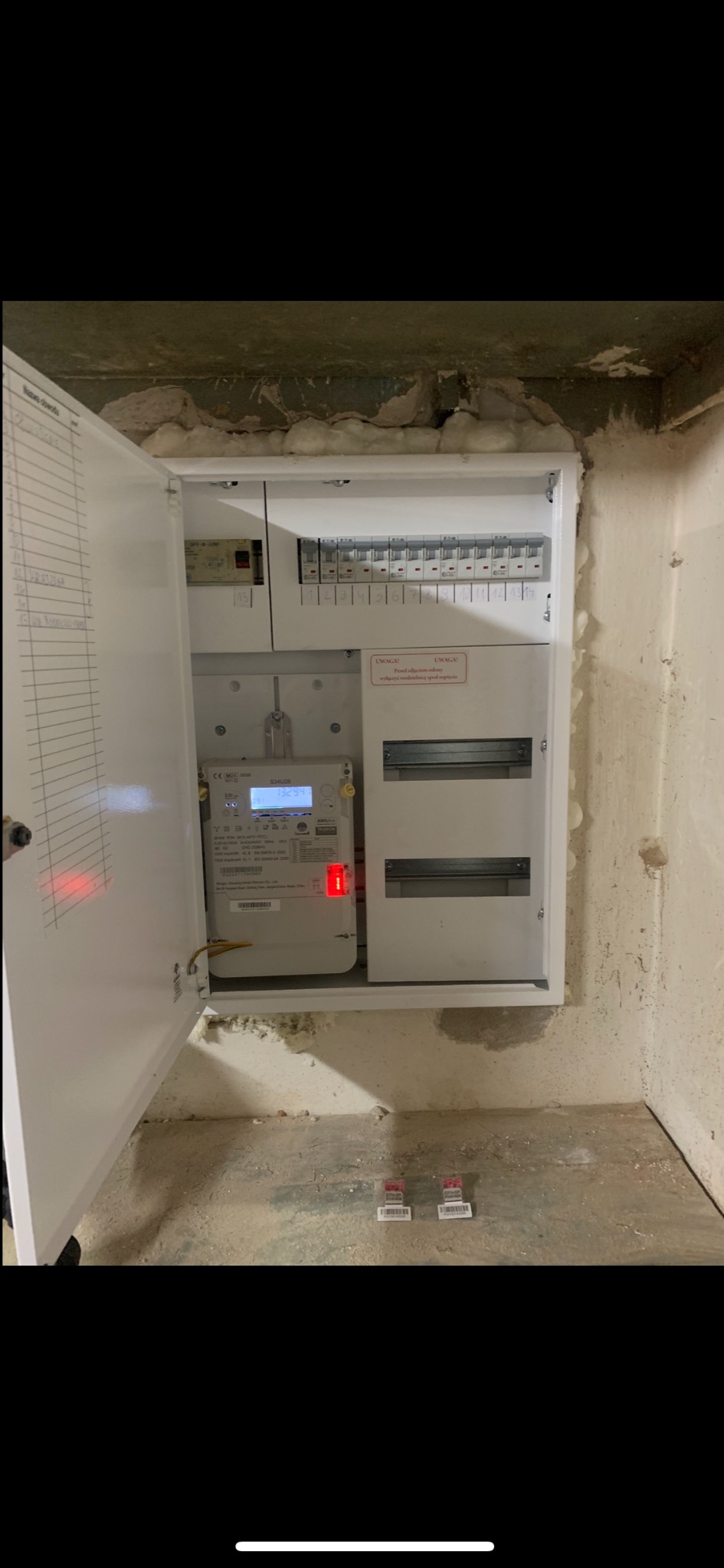 Otwarta skrzynka elektryczna z licznikiem i bezpiecznikami w surowym wnętrzu. Widoczne przewody i elementy instalacji. Na podłodze leżą etykiety.