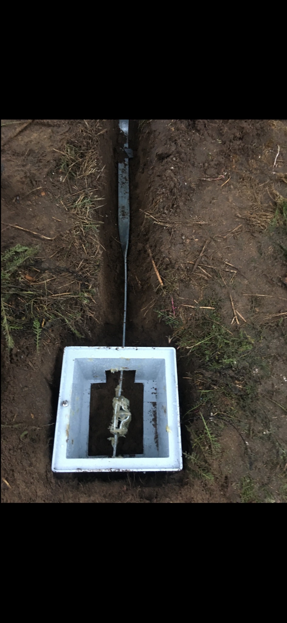 Uziemienie w wykopie z widoczną skrzynką kontrolną i metalową szyną, otoczone ziemią i roślinnością. Widok z góry na instalację uziemiającą.