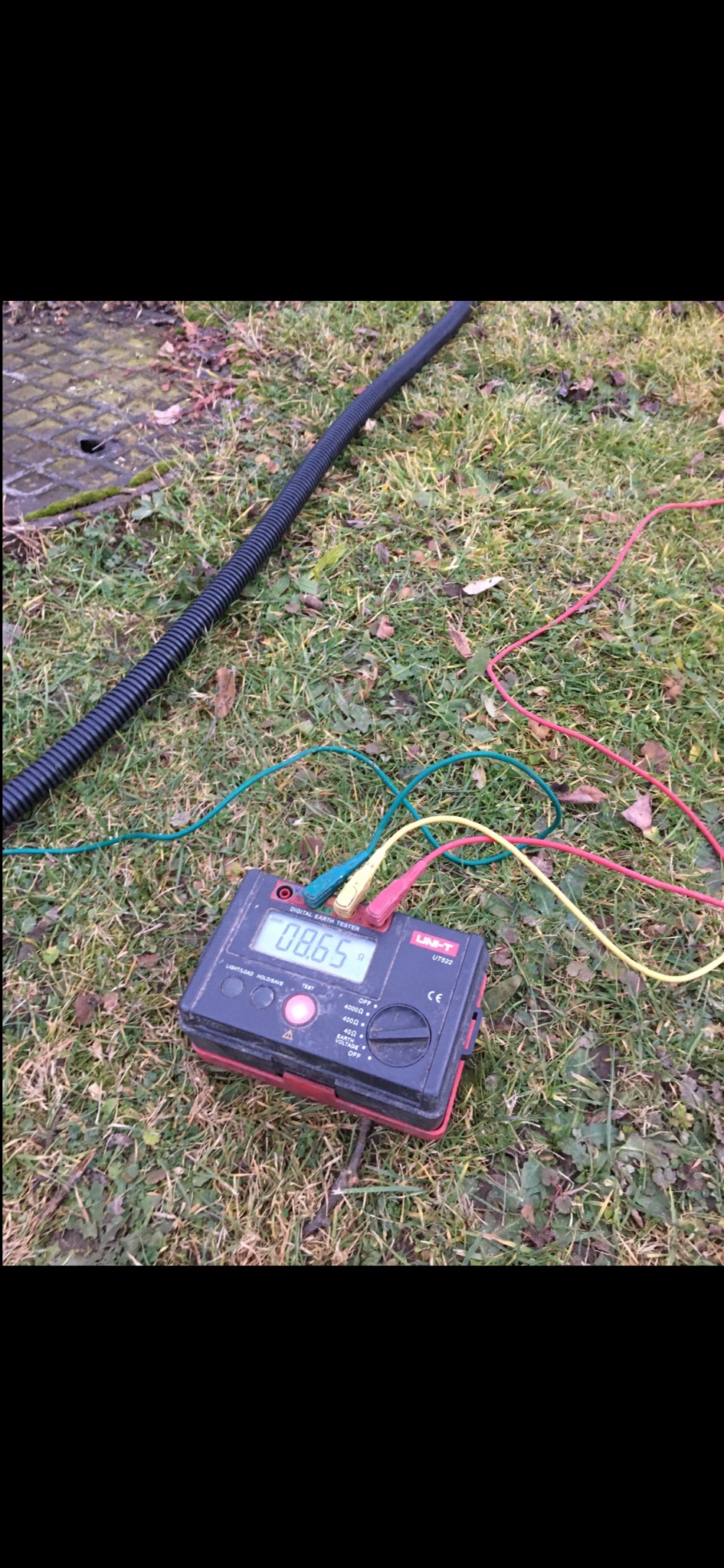 Miernik rezystancji uziomu Uni-T UT522 na trawie, z podłączonymi przewodami pomiarowymi. W tle czarna rura karbowana i studzienka kanalizacyjna.