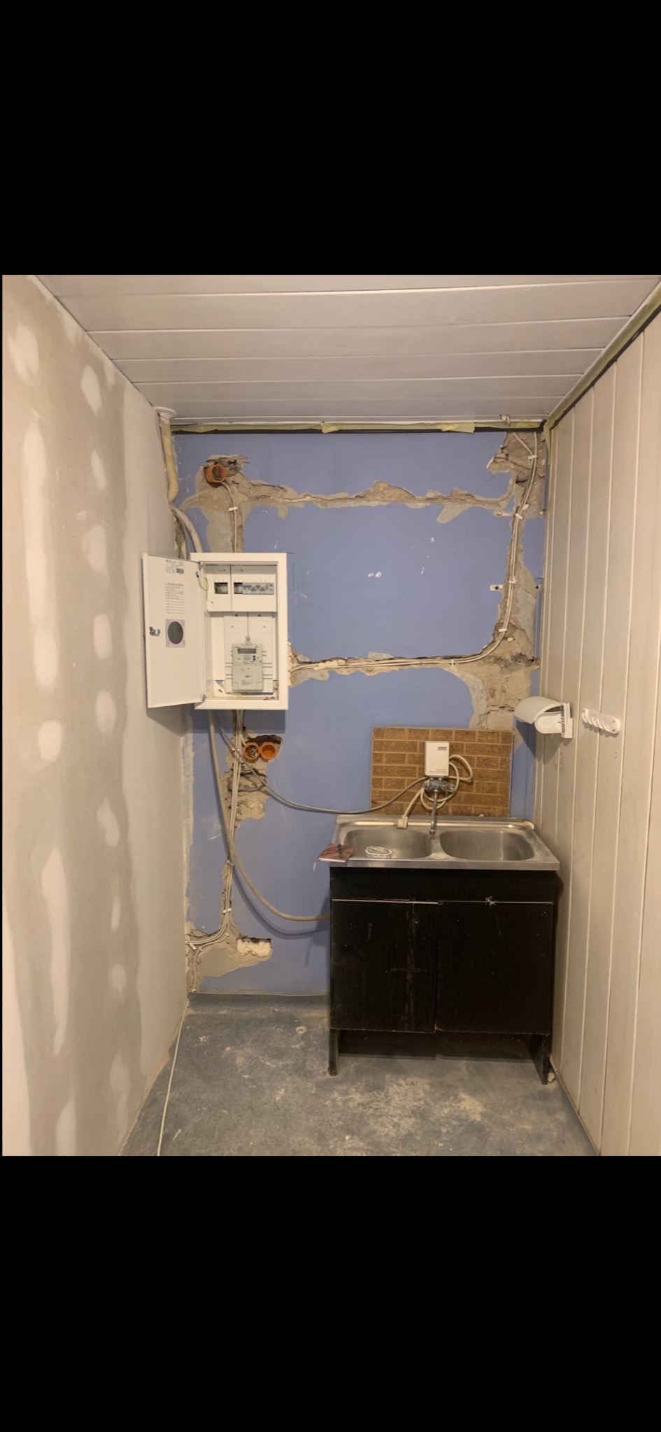 Otwarta skrzynka elektryczna z przewodami na niebieskiej ścianie w trakcie remontu. Widoczne kable, rury i fragmenty instalacji. Ściany częściowo otynkowane.