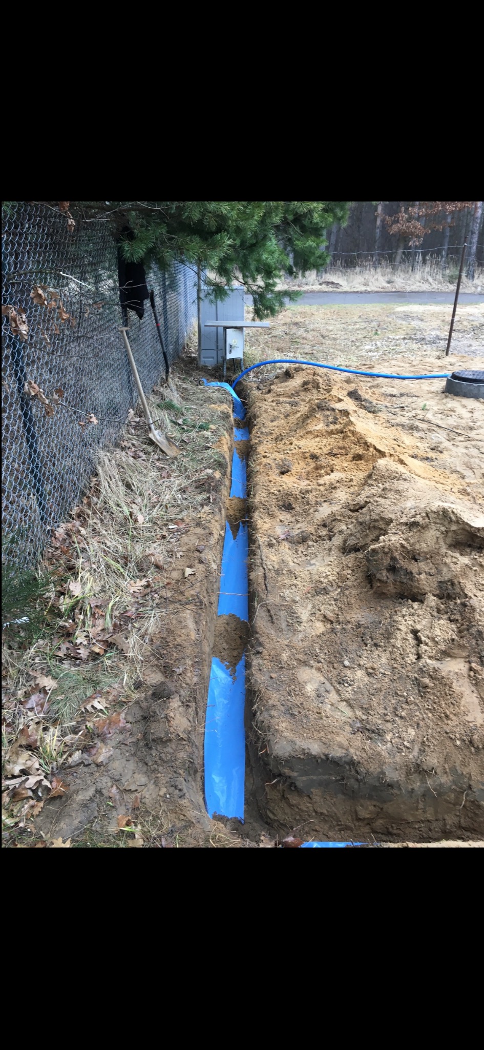 Wykop z niebieską rurą w ziemi, widoczny fragment instalacji przy ogrodzeniu z siatki. Szpadel wbity w ziemię obok wykopu. Etap prac instalacyjnych.
