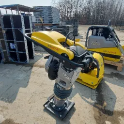 Żółto-szara ubijarka Wacker Neuson BS60-2 stoi na betonowym placu, w tle widoczna płyta wibracyjna DPU6555, również Wacker Neuson, oraz składowane elementy szalunkowe.