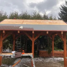 Montaż brązowego gontu bitumicznego na drewnianej altanie ogrodowej, widoczne narzędzia, drabina, częściowo ułożone deskowanie, w tle iglaki i pochmurne niebo.