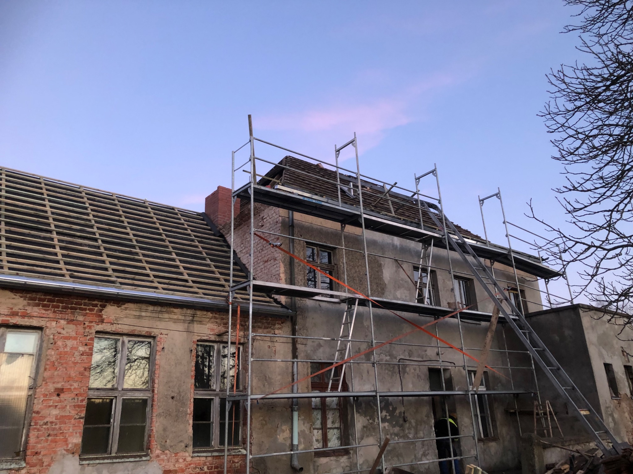 Budynek w trakcie renowacji dachu, widoczne rusztowania, częściowo odsłonięta konstrukcja dachu, stare okna i ceglana elewacja w tle błękitne niebo o zmierzchu.