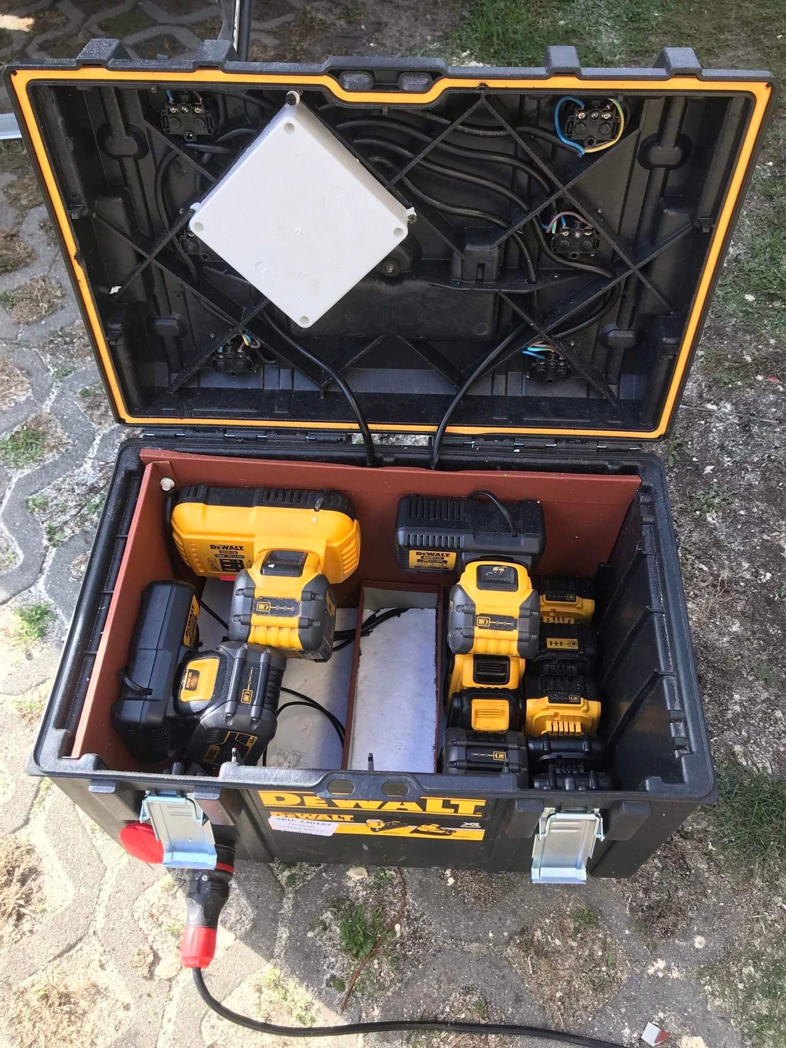 Otwarta skrzynka narzędziowa DeWalt z widocznymi narzędziami akumulatorowymi, ładowarkami i okablowaniem w górnej części pokrywy, zasilana kablem z czerwoną wtyczką.