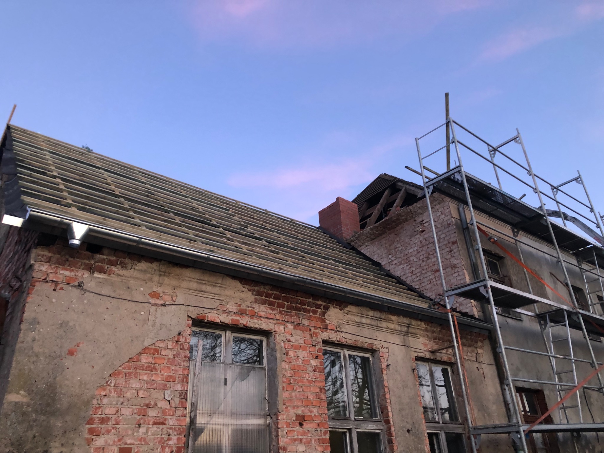 Remontowany budynek z odsłoniętą więźbą dachową, widoczne rusztowania i fragmenty muru z cegły.