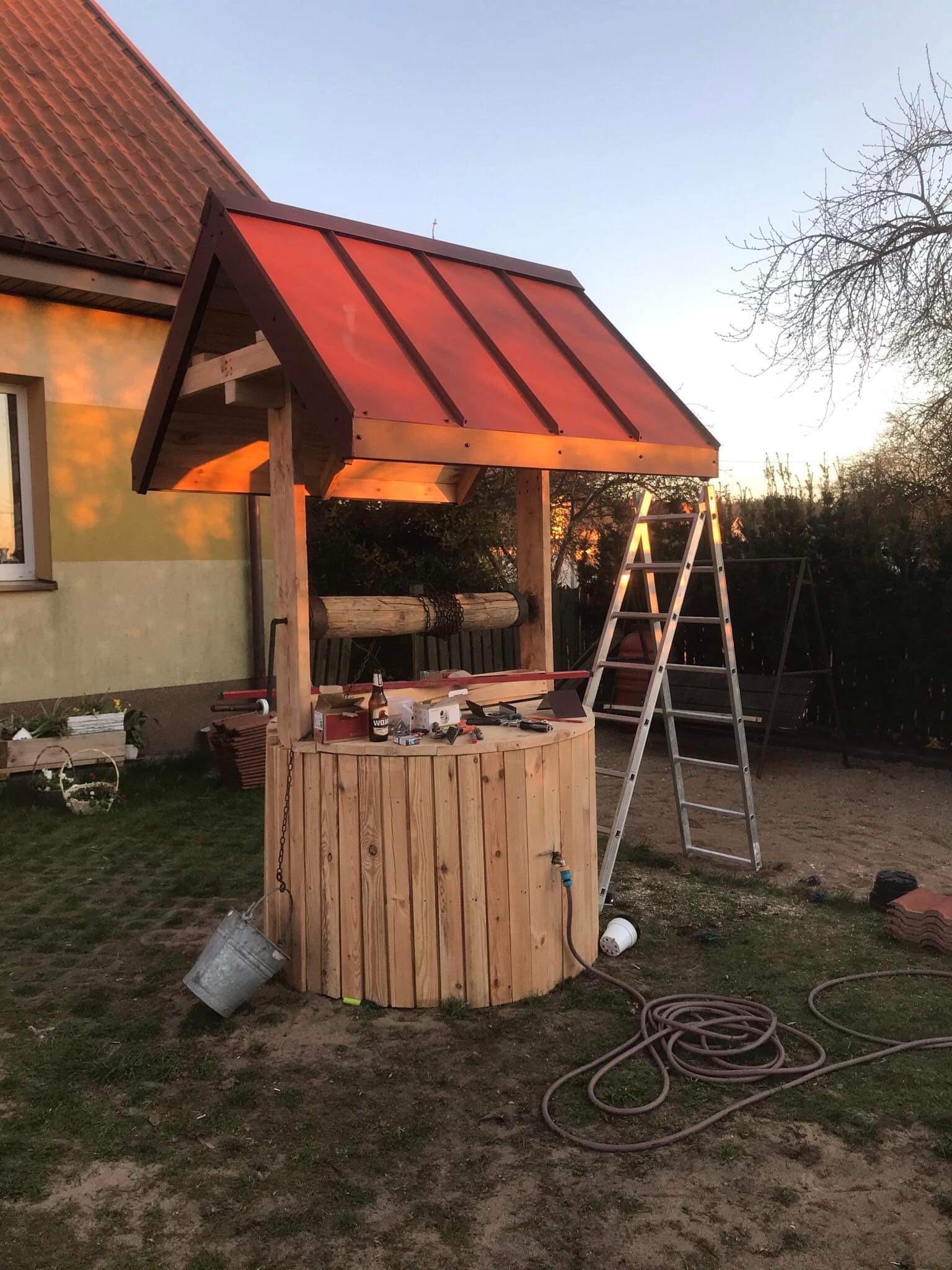 Konstrukcja studni z drewnianą obudową i czerwonym dachem w trakcie budowy, widoczna drabina, narzędzia i materiały budowlane w otoczeniu trawnika i domu.
