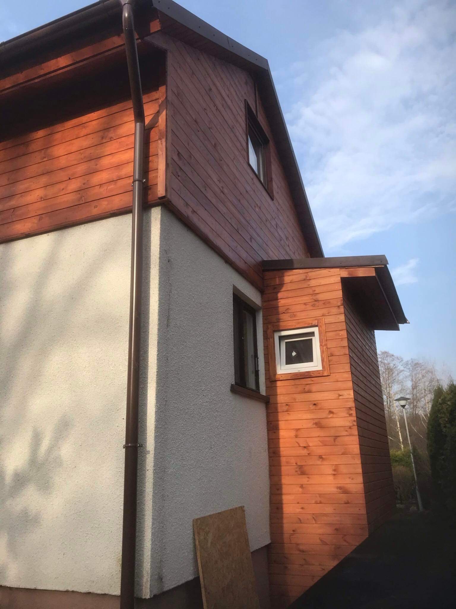 Dom jednorodzinny z brązową drewnianą elewacją, widoczny system rynnowy w kolorze brązowym, fragment otynkowanej ściany oraz skrawek płyty OSB przy ścianie.