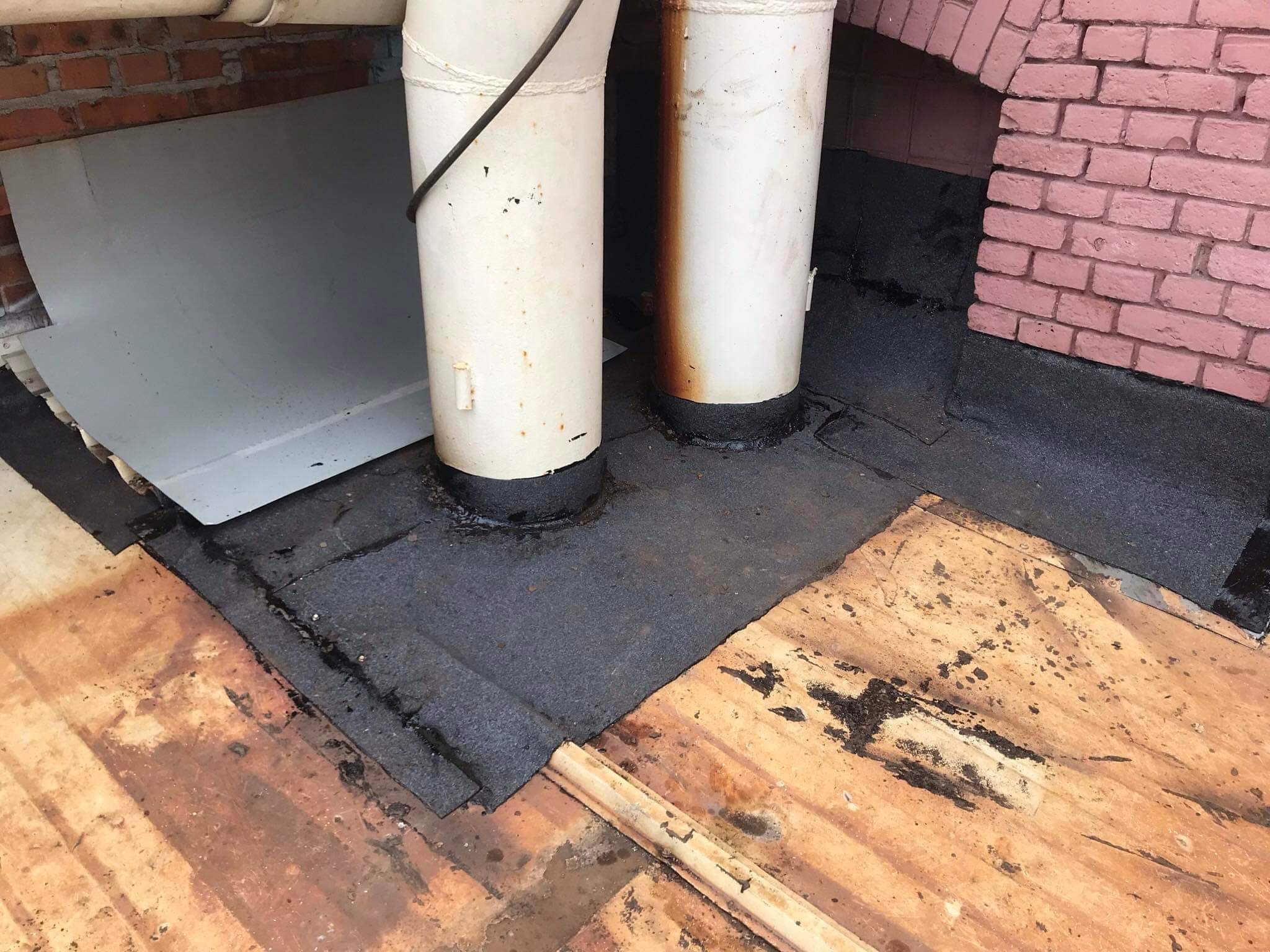 Fragment dachu z dwoma pionowymi rurami, zabezpieczonymi czarną, bitumiczną izolacją, przylegający do ceglanego komina, z widocznym fragmentem blachy i śladami wilgoci na drewnianym podłożu.