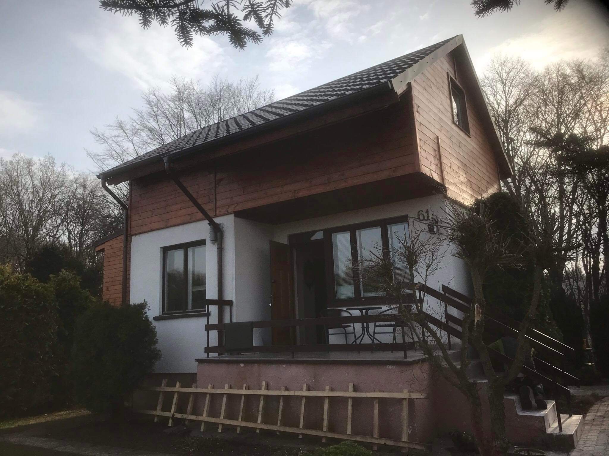 Dwukondygnacyjny dom z ciemnobrązowym dachem z dachówki bitumicznej, drewnianą elewacją na piętrze i białym tynkiem na parterze, widoczny fragment rynny i drabina oparta o fundament.