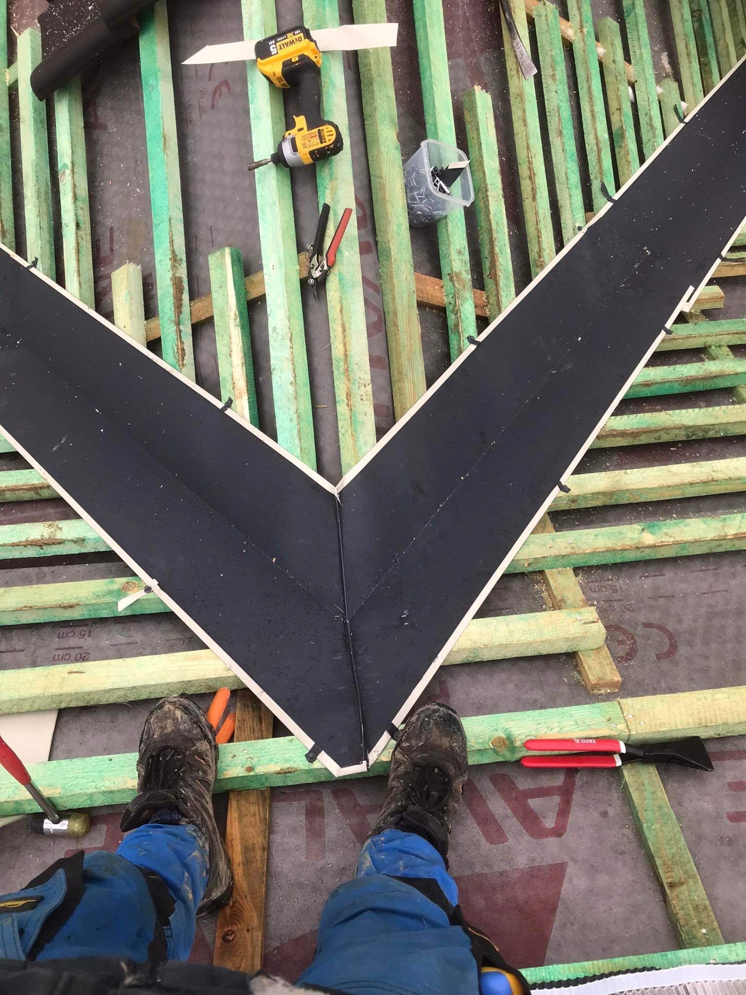 Montaż obróbki blacharskiej w kształcie litery V na dachu w Gorzeniu, widok z góry na konstrukcję z łat i kontrłat, narzędzia dekarskie i buty robocze.