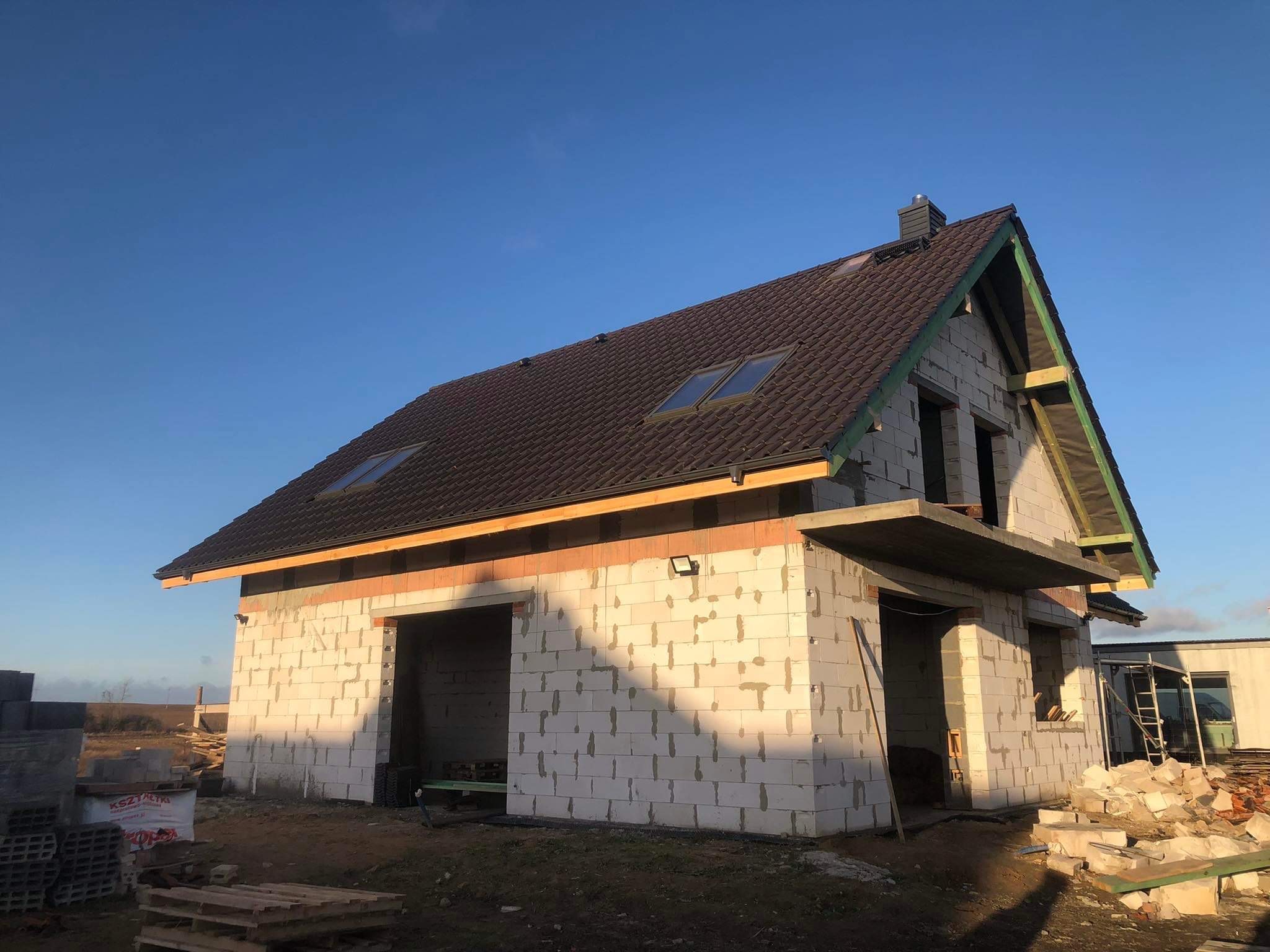 Dom w stanie surowym zamkniętym z brązową dachówką, widoczne okna dachowe i przygotowane otwory okienne, ściany z białego bloczka, na pierwszym planie palety z materiałami budowlanymi.