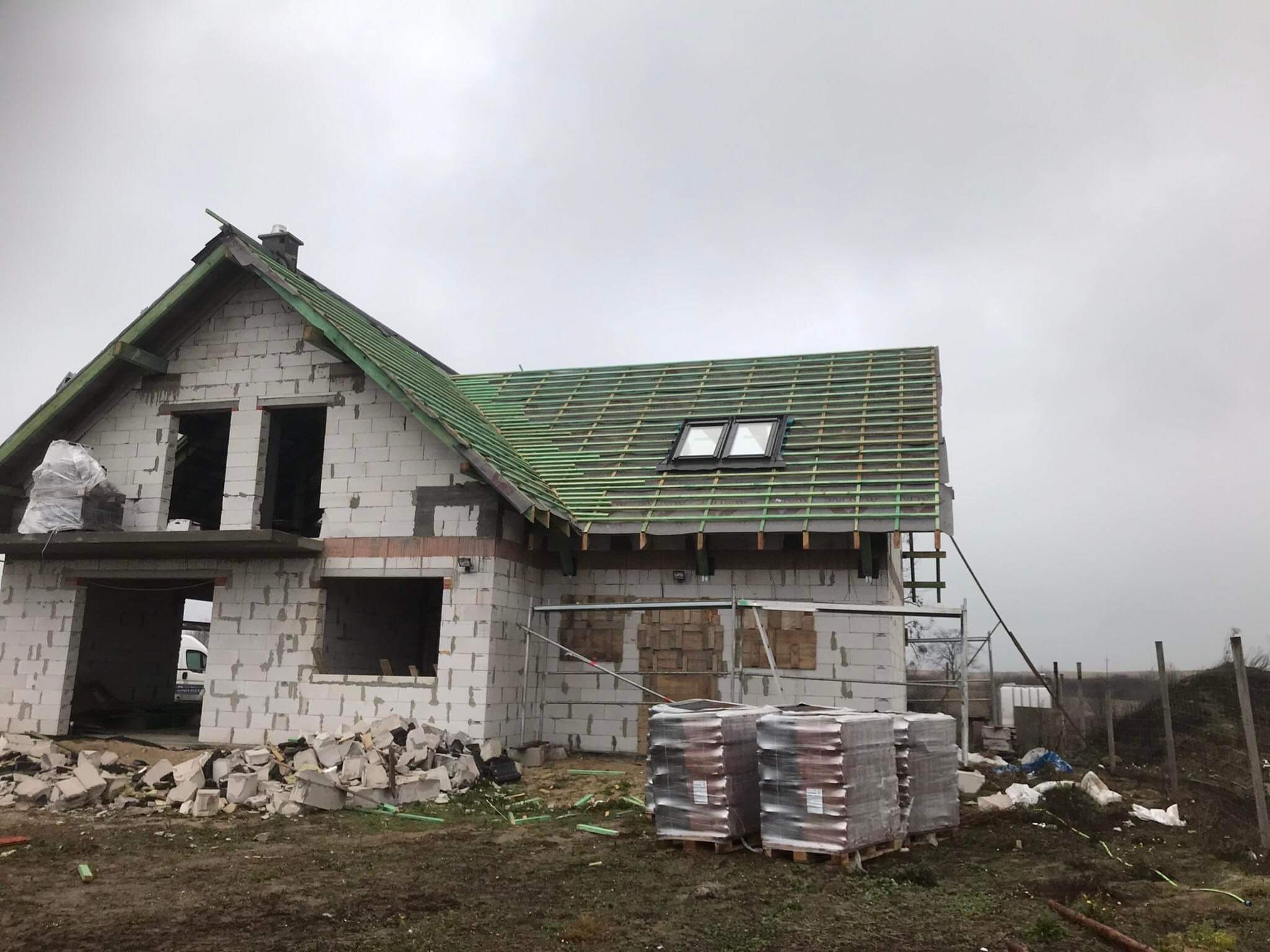 Dom w trakcie budowy z widoczną konstrukcją dachu pokrytą zieloną membraną i oknami dachowymi, palety z materiałami budowlanymi na pierwszym planie, gruz i resztki materiałów wokół.
