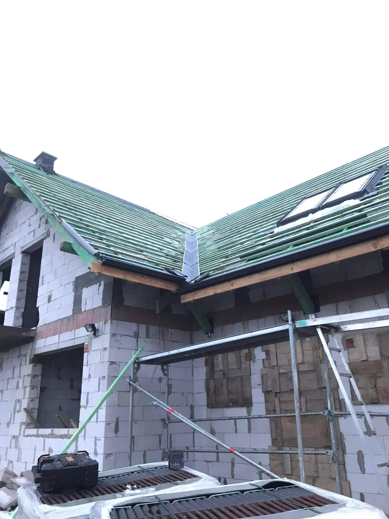 Dom w trakcie budowy z widoczną konstrukcją dachu z łatami, oknami dachowymi i przygotowanymi obróbkami blacharskimi, z rusztowaniem i narzędziami na pierwszym planie.