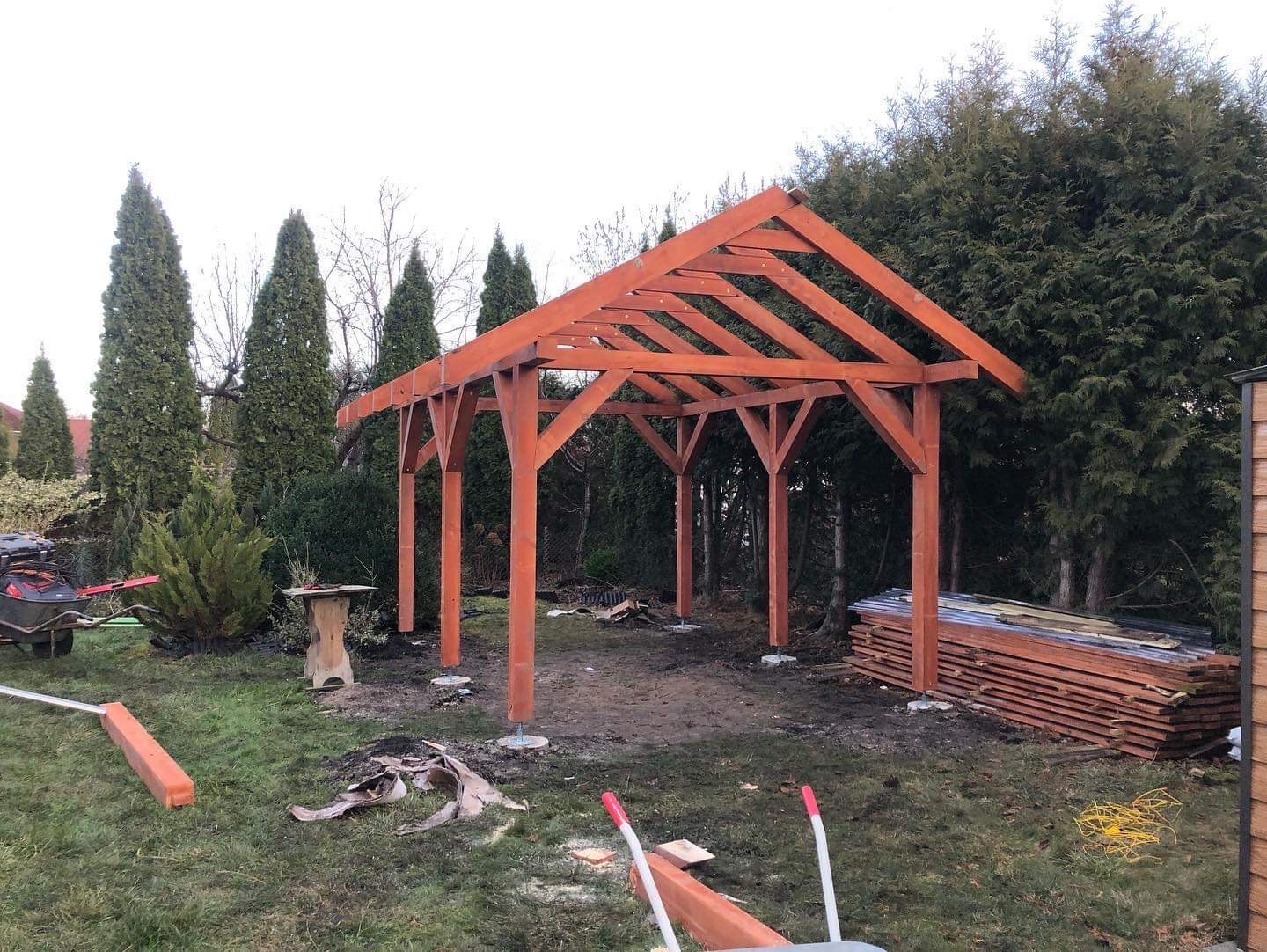 Konstrukcja drewniana altany w trakcie budowy, widoczne słupy i więźba dachowa w kolorze pomarańczowym, na trawie leżą resztki materiałów i narzędzia, w tle wysokie tuje.