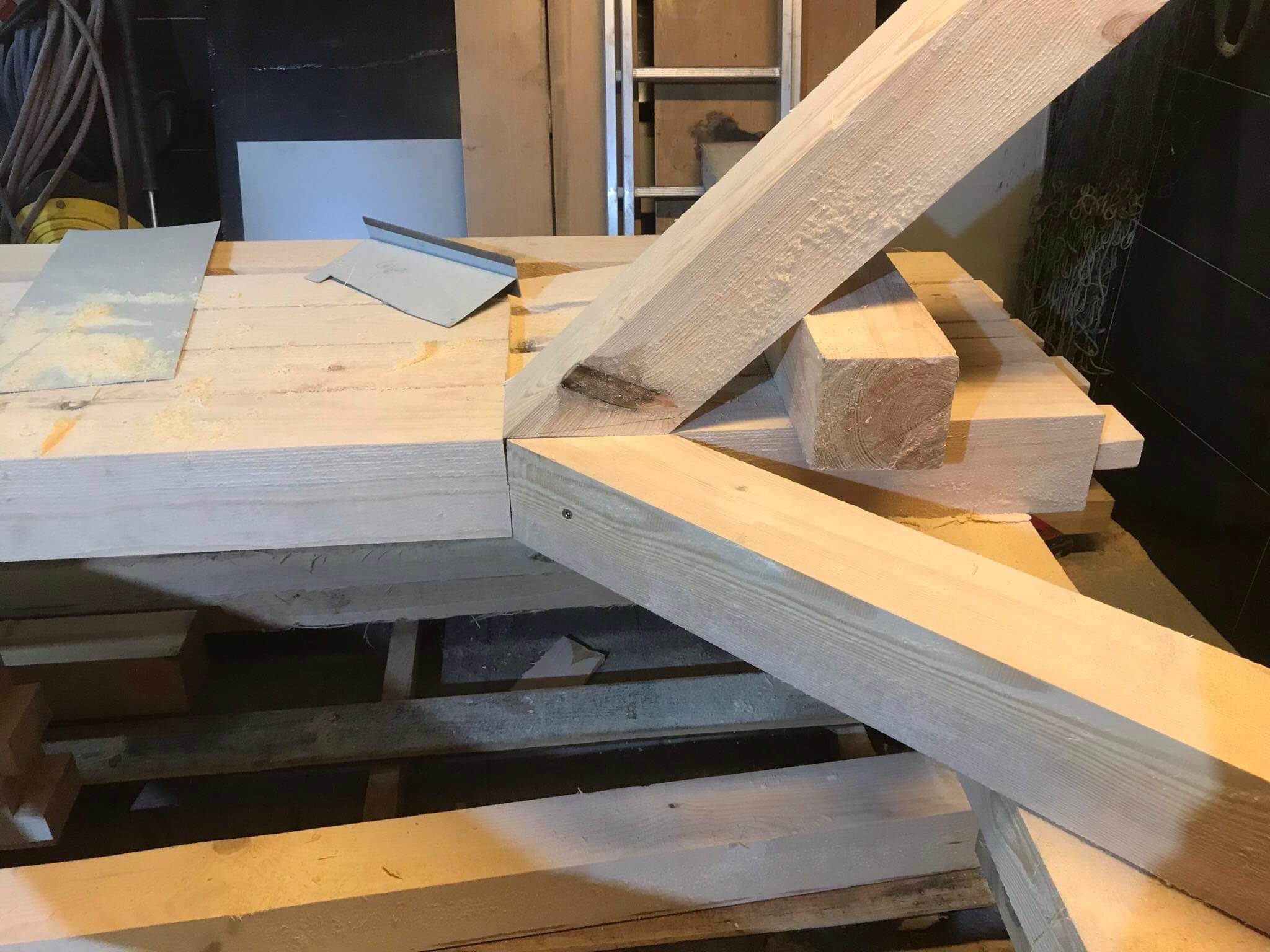 Fragment konstrukcji drewnianej w warsztacie, elementy więźby dachowej z widocznymi śladami obróbki i metalowymi elementami mocującymi, drabina w tle.