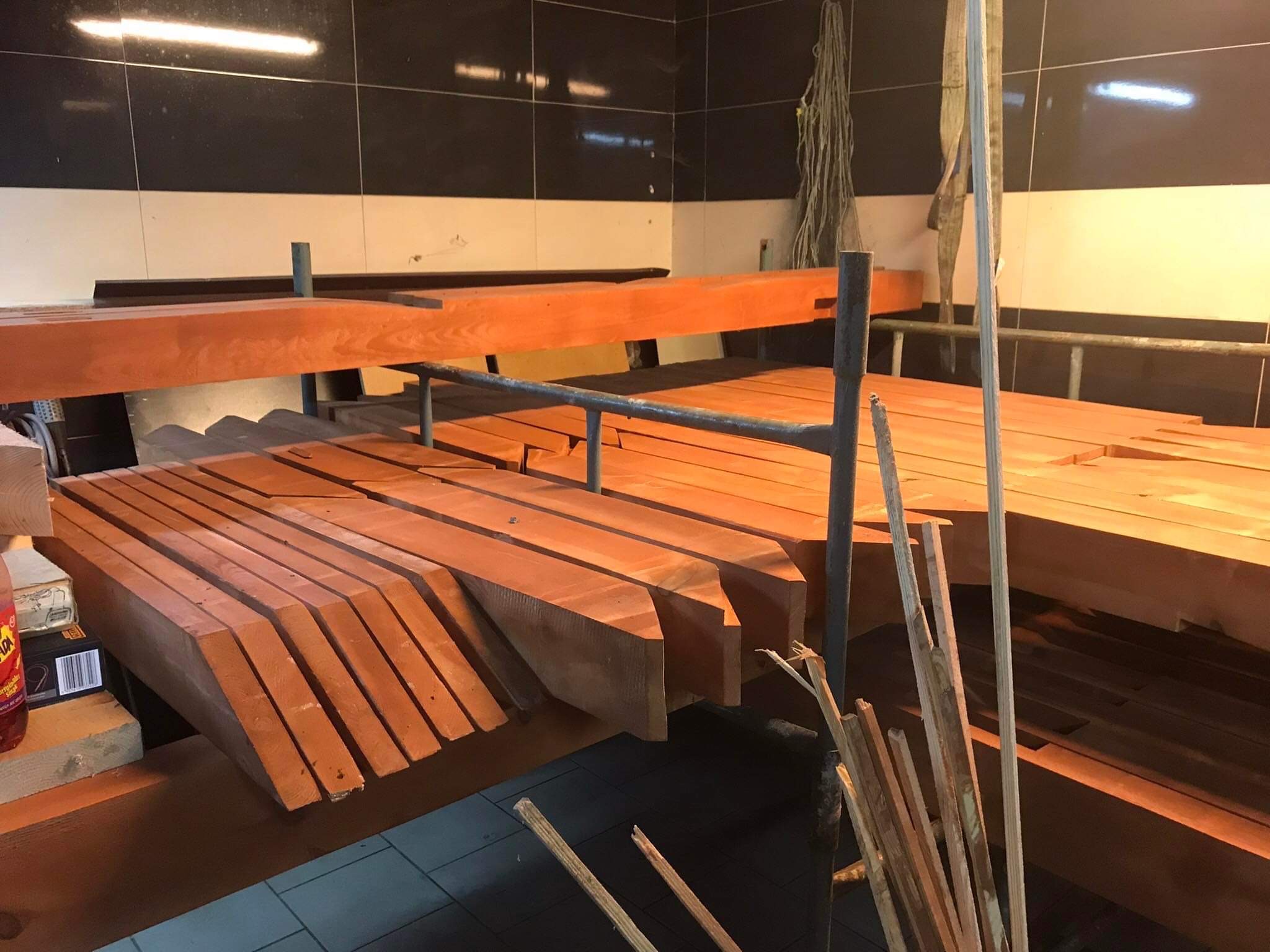 Przygotowane drewniane elementy konstrukcyjne, prawdopodobnie krokwie dachowe, ułożone na rusztowaniu w pomieszczeniu z czarno-białymi płytkami na ścianach. Widoczne odpady drewniane i napój w tle.