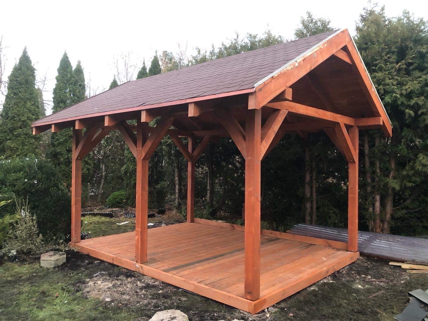 Drewniana altana z brązowym dachem z gontu bitumicznego w ogrodzie, widok z zewnątrz na konstrukcję nośną i podłogę z desek.