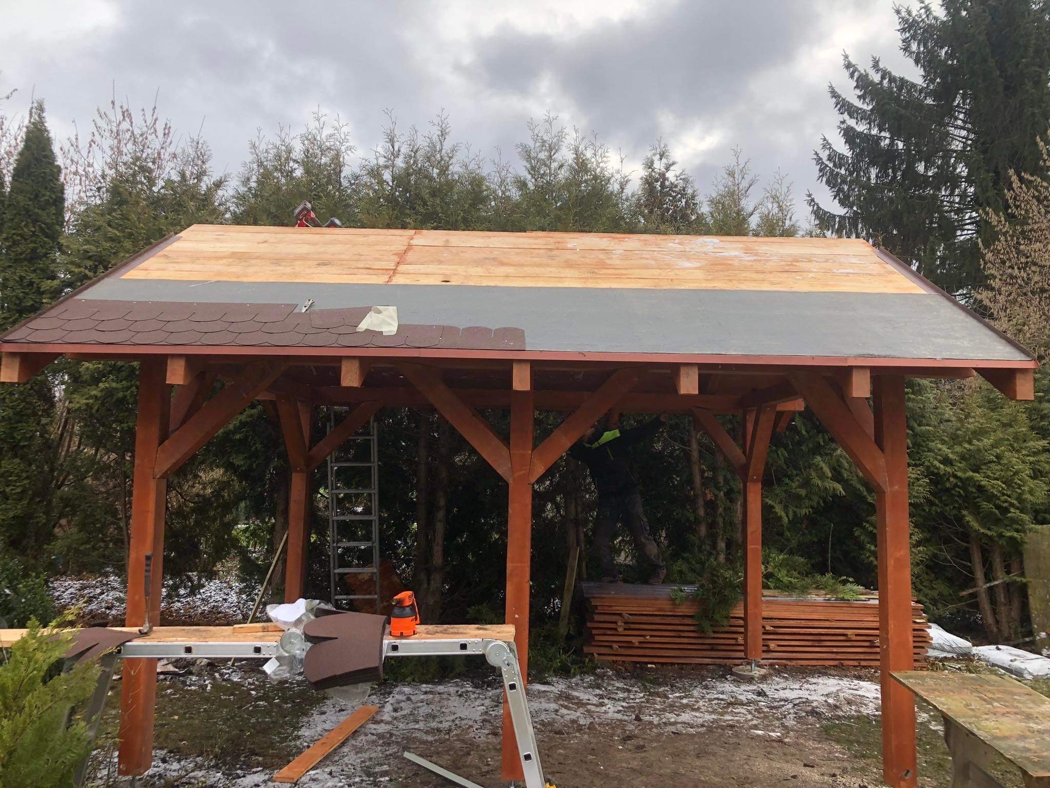 Montaż brązowego gontu bitumicznego na drewnianej altanie ogrodowej, widoczne narzędzia, drabina, częściowo ułożone deskowanie, w tle iglaki i pochmurne niebo.