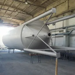 Pomalowany na jasnoszaro przemysłowy zbiornik ciśnieniowy na stalowych podporach w hali produkcyjnej, widoczne rury wentylacyjne i drabina.