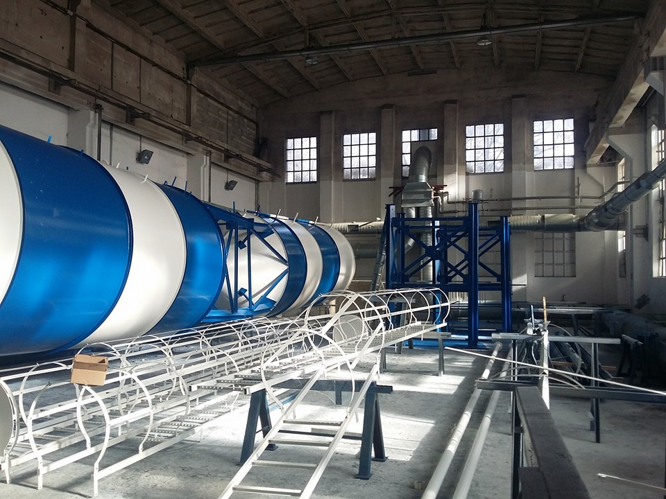 Wnętrze hali przemysłowej z dużym, cylindrycznym elementem w kolorach białym i niebieskim oraz metalową konstrukcją rusztowania na pierwszym planie.