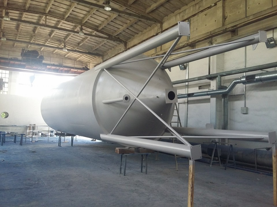 Pomalowany na jasnoszaro przemysłowy zbiornik ciśnieniowy na stalowych podporach w hali produkcyjnej, widoczne rury wentylacyjne i drabina.