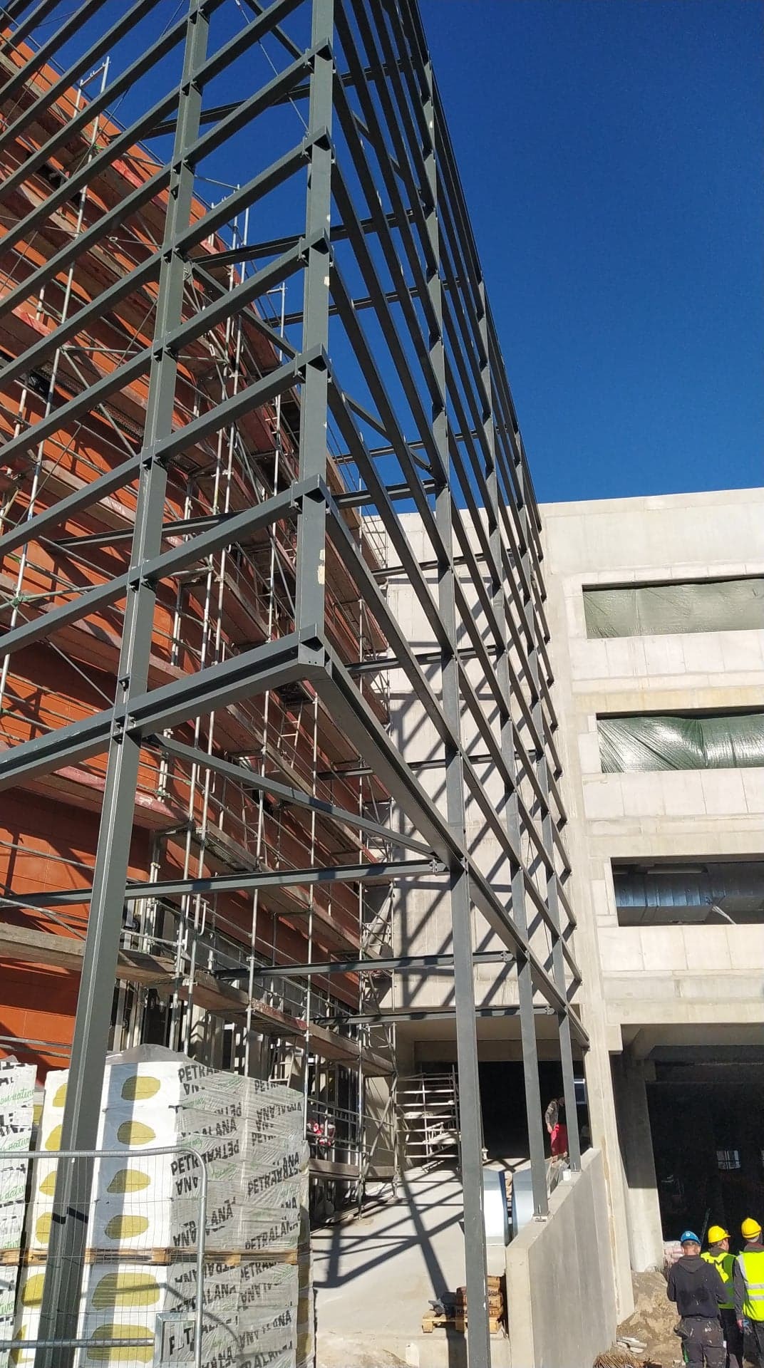 Konstrukcja stalowa budynku w trakcie montażu, widoczne rusztowania, materiały budowlane i pracownicy w żółtych kamizelkach na tle błękitnego nieba.