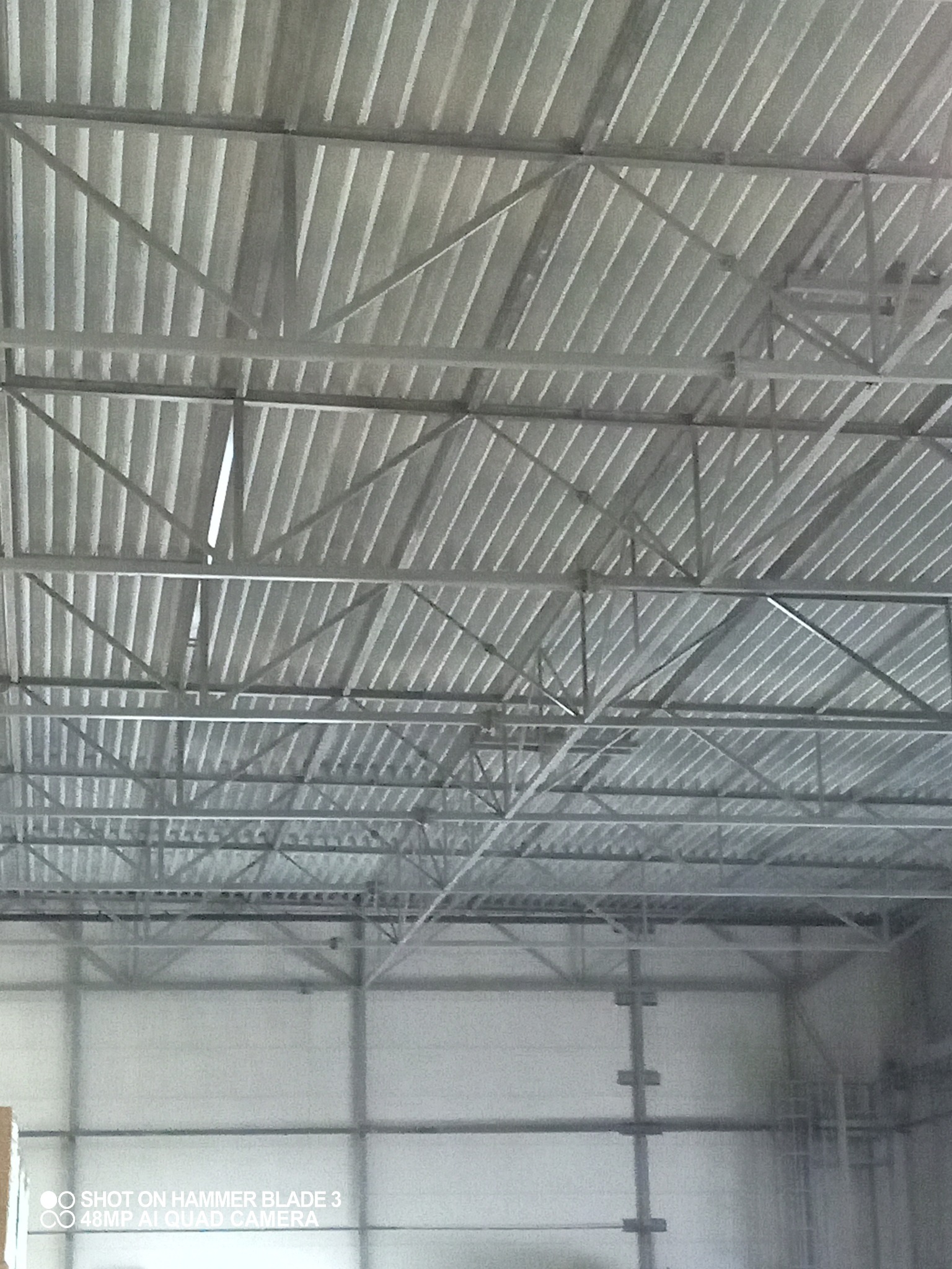 Wnętrze hali magazynowej z widoczną konstrukcją stalową dachu pokrytą blachą trapezową, oświetloną naturalnym światłem wpadającym przez szczeliny.