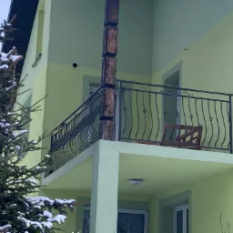 Balkon z czarną, kutą balustradą, drewnianym słupem z bali, widok na krzesła i stół, jasna elewacja budynku w tle, częściowo zasłonięty przez ośnieżone drzewo iglaste.