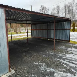 Zadaszona wiata garażowa z blachy falistej, wsparta na metalowych słupach, na żwirowym podłożu z resztkami śniegu, widok na drzewa w tle.