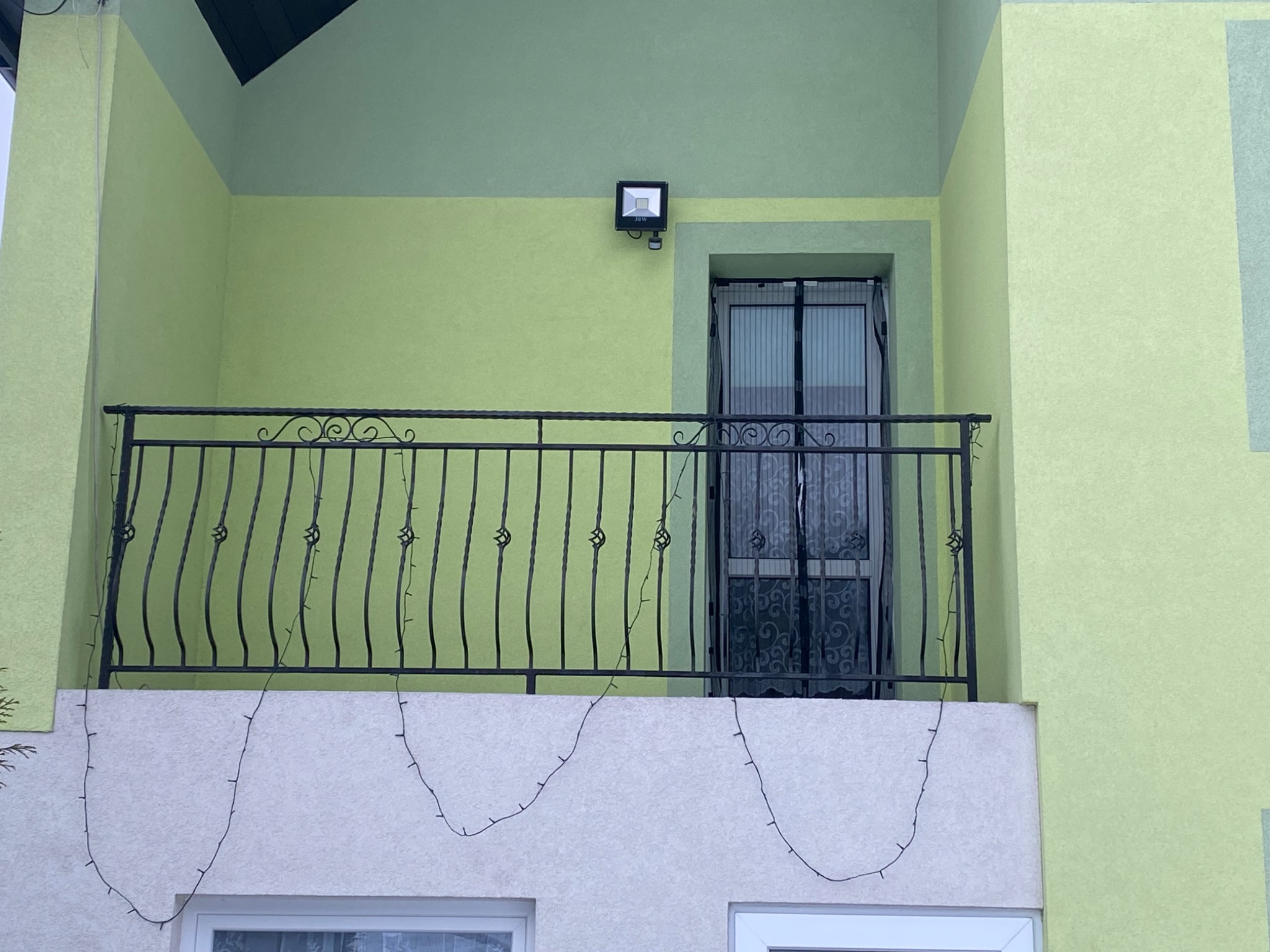 Kuty balkon z czarną, ozdobną balustradą, oświetlony lampą zewnętrzną, dekorowany wiszącymi lampkami, na tle jasnozielonej elewacji budynku.