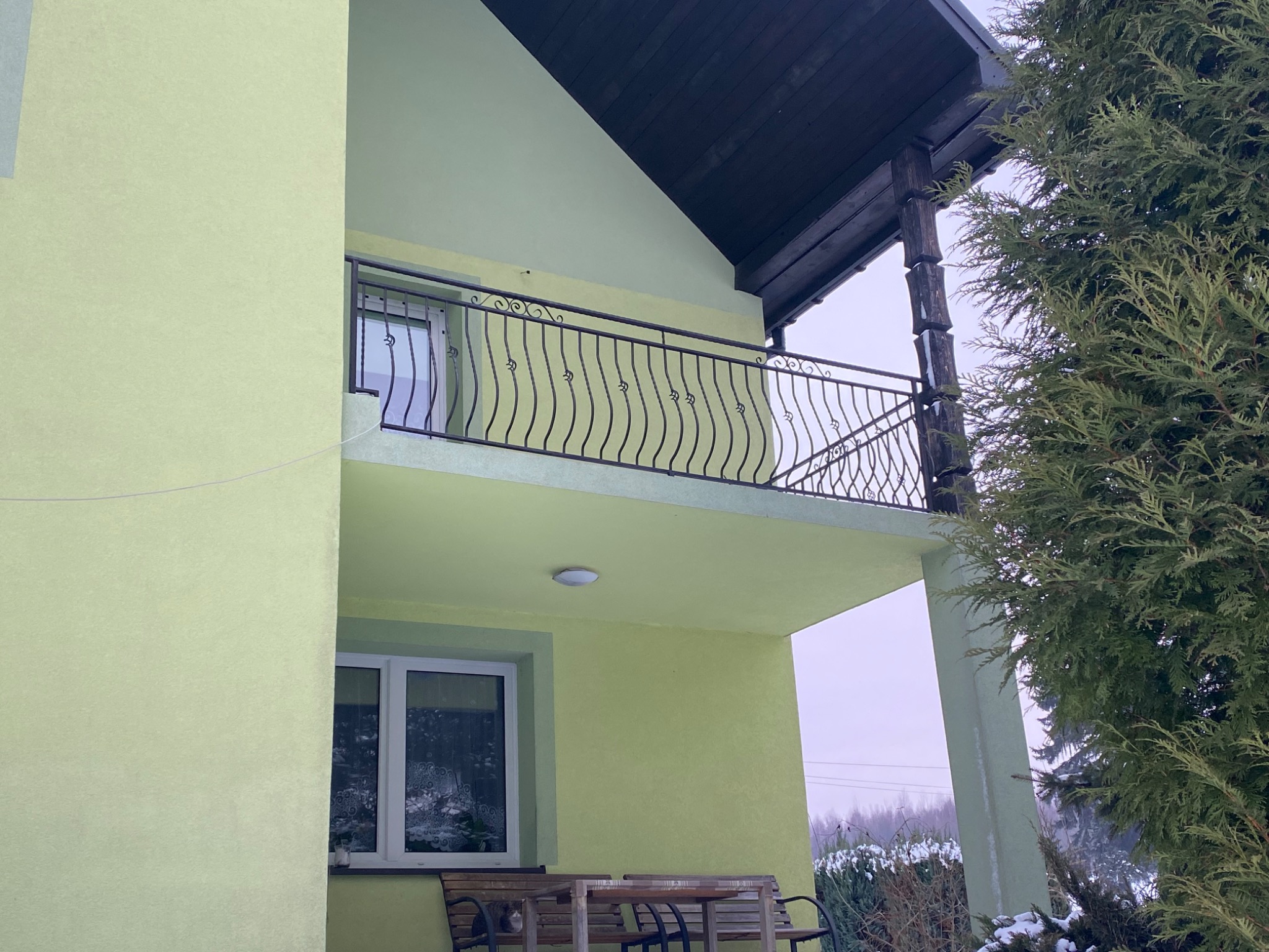 Żółto-zielony dom z balkonem z czarną, ozdobną balustradą, widok z dołu, z fragmentem ogrodu i drzewa iglastego.