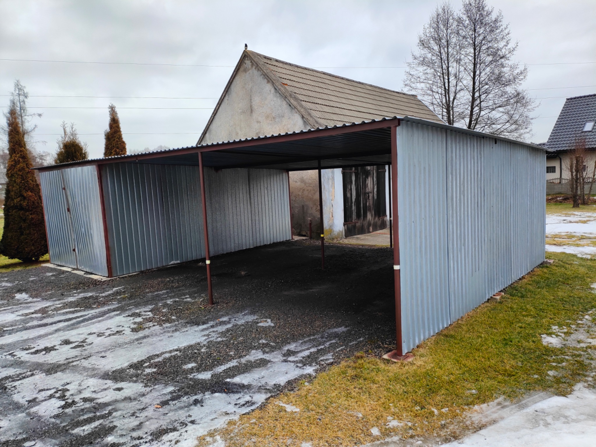 Zadaszone stanowisko parkingowe z blachy falistej na posesji w pochmurny dzień, widoczne pozostałości śniegu na podłożu.