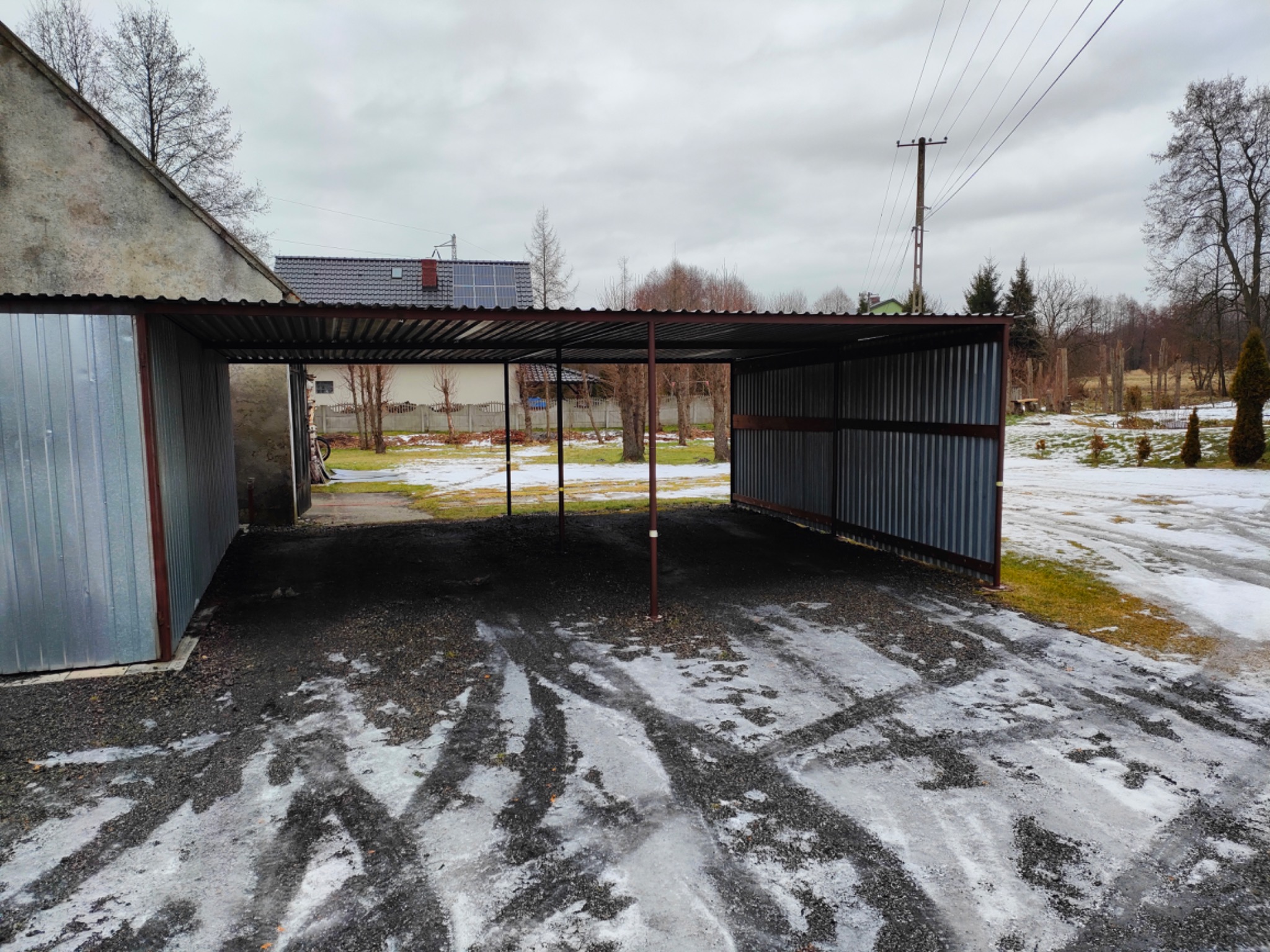 Zadaszona wiata garażowa z metalową konstrukcją i falistymi blachami, na żwirowym podłożu z resztkami śniegu, widok z zewnątrz w pochmurny dzień.