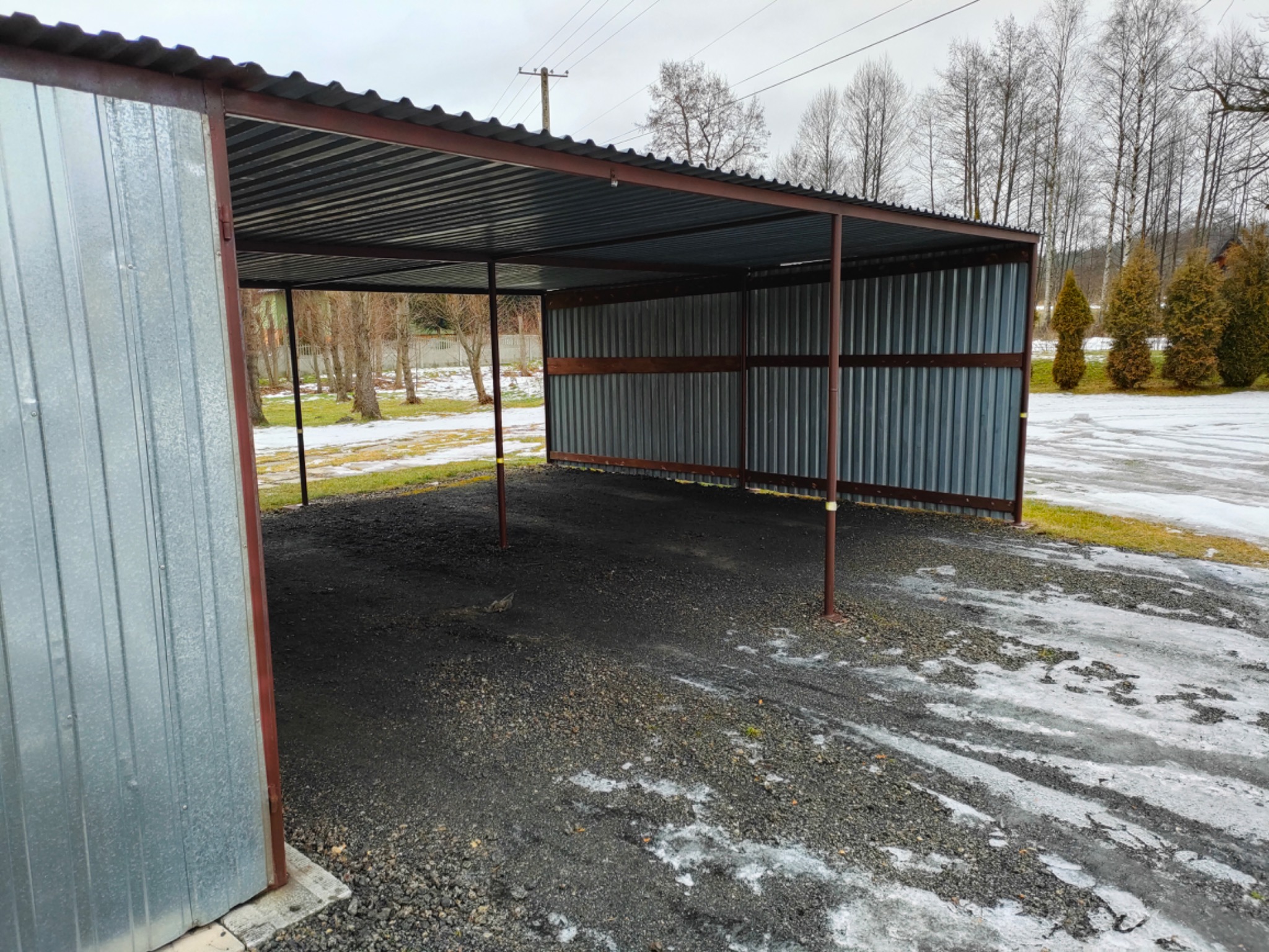 Zadaszona wiata garażowa z blachy falistej, wsparta na metalowych słupach, na żwirowym podłożu z resztkami śniegu, widok na drzewa w tle.