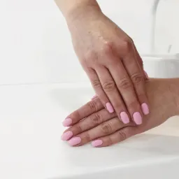 Dłonie kobiety z świeżo pomalowanymi, krótkimi paznokciami w odcieniu pastelowego różu, ułożone na białym blacie w salonie kosmetycznym.