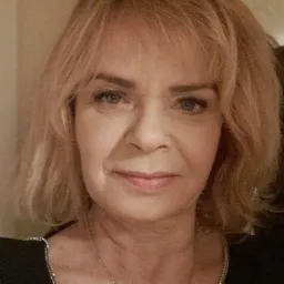 Portret kobiety o blond włosach i niebieskich oczach, uśmiechająca się subtelnie, ubrana w czarną bluzkę z srebrnym wykończeniem dekoltu i delikatny srebrny łańcuszek.