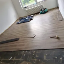 Fragment pokoju z częściowo ułożoną podłogą z paneli w odcieniach brązu, widoczne narzędzia i paczki paneli, stare podłoże w dolnej części kadru.